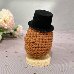 May include: A handmade, crocheted figure in shades of brown, wearing a small black top hat. The figure is mounted on a round, wooden base. The background features pink and gold decorative elements.