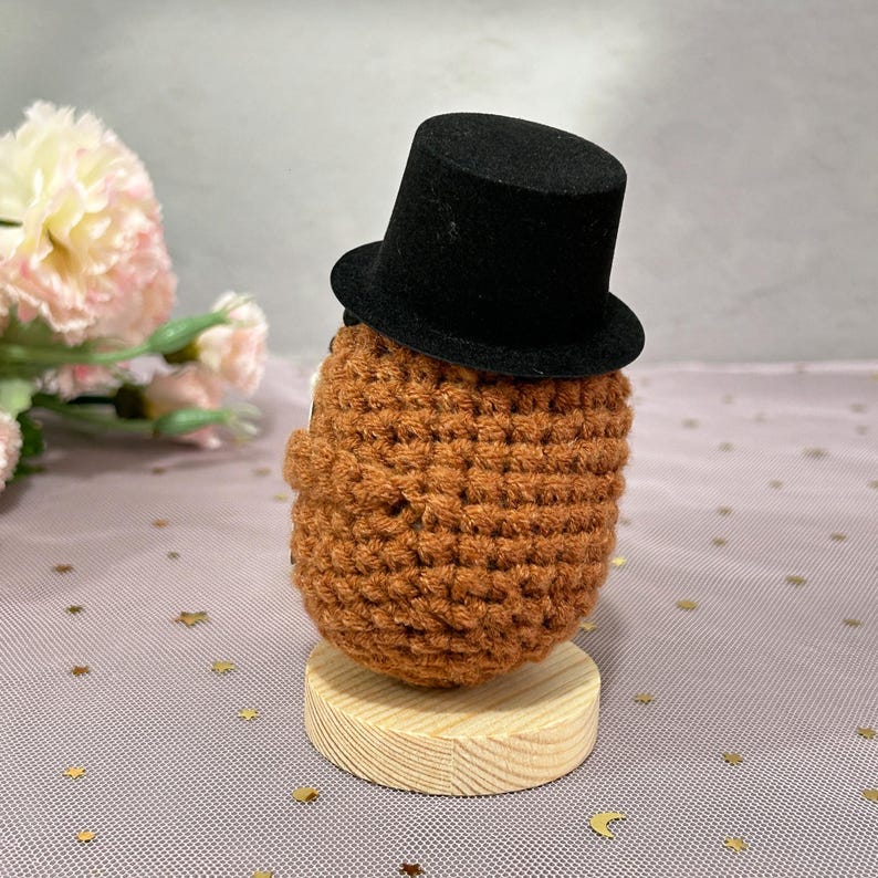 May include: A handmade, crocheted figure in shades of brown, wearing a small black top hat. The figure is mounted on a round, wooden base. The background features pink and gold decorative elements.