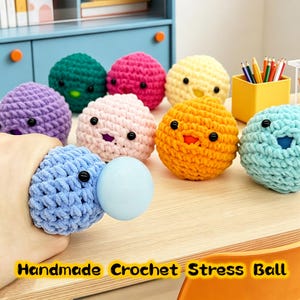 Puede incluir: Una colección de bolas antiestrés hechas a mano con ganchillo en varios colores, incluyendo azul, morado, rosa, verde, amarillo y naranja. Cada bola tiene una cara con ojos negros y boca. Una bola azul está siendo apretada, revelando una bola azul claro.