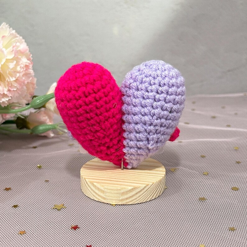 May include: A handmade crochet heart, split into two halves, one bright pink and the other lavender. The heart is mounted on a small, round wooden base. The craft project is a decorative item.