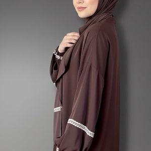 May include: A brown abaya with white embroidered leaf patterns on the sleeves and front. The abaya is worn with a matching brown hijab. The sleeves are long and loose, and the fabric appears to be soft and flowing.