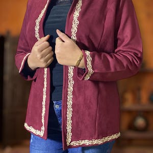 May include: A burgundy jacket with cream-coloured embroidered trim. The jacket has a tailored fit and is open in the front. The trim is along the edges of the jacket and sleeves. The jacket is worn over a black top and blue jeans.