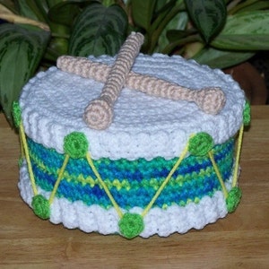 Puede incluir: Un tambor de juguete de crochet con rayas verdes y azules y ribete blanco. El tambor tiene dos baquetas de crochet blancas.