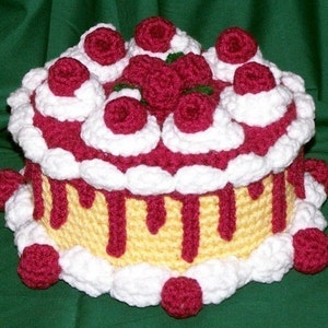 May include: A crocheted cake with white frosting and red cherries. The cake is decorated with red icing and has a yellow base.