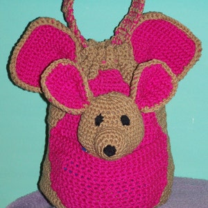 Puede incluir: Una bolsa de ratón de crochet rosa y marrón con cierre de cordón. La bolsa tiene orejas grandes y una nariz pequeña.