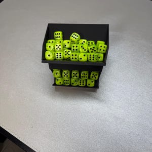 May include: A two-tiered black plastic storage rack filled with numerous neon yellow dice. Each die features black pips on all sides. The rack is designed with open-front compartments that hold the dice in organized rows, displaying a bright contrast between the black structure and the vibrant yellow game pieces.