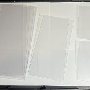 May include: A collection of clear, rectangular plastic sheets, likely for protective packaging or crafting. The sheets are arranged in stacks and individually, varying in size and quantity, against a white background.
