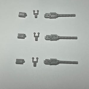 May include: Three sets of small, gray plastic model parts are arranged on a white surface. Each set includes a rectangular piece, a Y-shaped piece, and a long, rectangular piece with detailed features, resembling miniature weaponry.