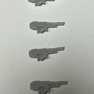 May include: Four gray plastic miniature gun models are arranged vertically against a light gray background. Each model features a rectangular body, a barrel, and a curved handle. The overall design suggests a futuristic or science fiction theme.