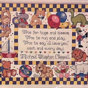 Peut inclure: Broderie au point de croix encadrée avec des jouets brodés et le texte "Time for hugs and kisses..." dans un motif en damier. Les jouets comprennent un voilier, un ours, un chien et un dinosaure.