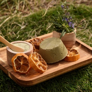 Peut inclure: Un plateau en bois avec un pain de shampooing vert, un petit bol de poudre verte avec une cuillère en bois et des tranches d'orange séchées. Un petit pot de romarin est en arrière-plan.