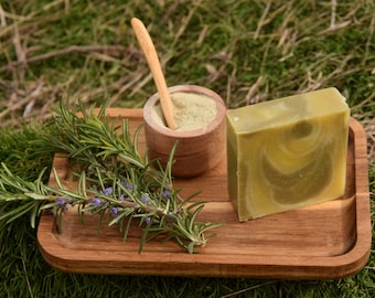 Può includere: Un vassoio di legno con una saponetta verde, una piccola ciotola di legno con polvere verde e un rametto di rosmarino con fiori viola.