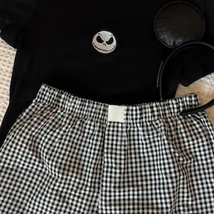 May include: A black t-shirt with a Jack Skellington face patch, black and white checked boxer shorts, and a black mouse ear headband. The shorts also feature a Jack Skellington face patch. This is likely a themed sleepwear set.