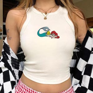 May include: White tank top featuring a colorful graphic of a mermaid and a yellow fish. The sleeveless top has a scoop neck and is made of a soft, comfortable material. The mermaid graphic is in shades of red, blue, and yellow.
