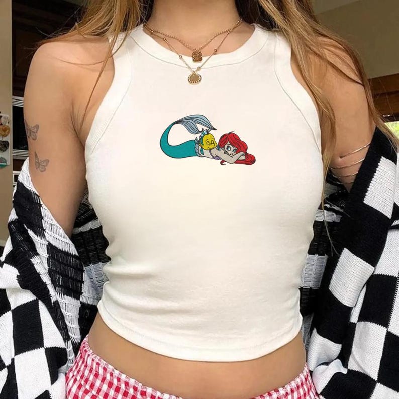 May include: White tank top featuring a colorful graphic of a mermaid and a yellow fish. The sleeveless top has a scoop neck and is made of a soft, comfortable material. The mermaid graphic is in shades of red, blue, and yellow.