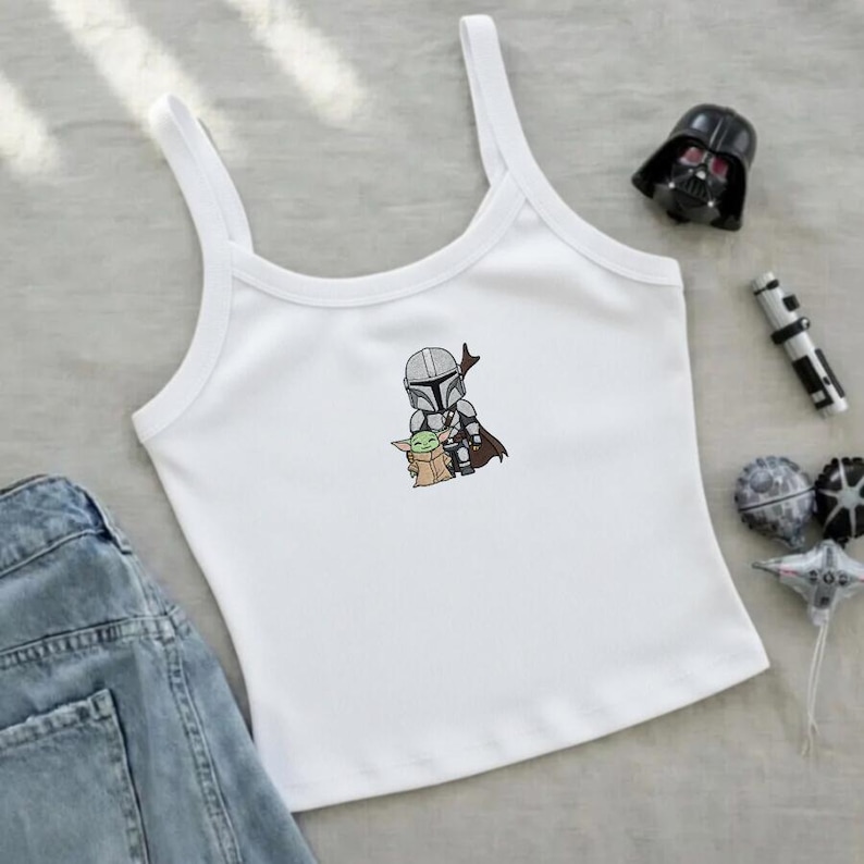 May include: White tank top with a small embroidered graphic of The Mandalorian and Grogu. The top has thin straps and a scoop neckline. A pair of blue denim shorts is visible on the left side of the image. Accessories include a Darth Vader figurine.