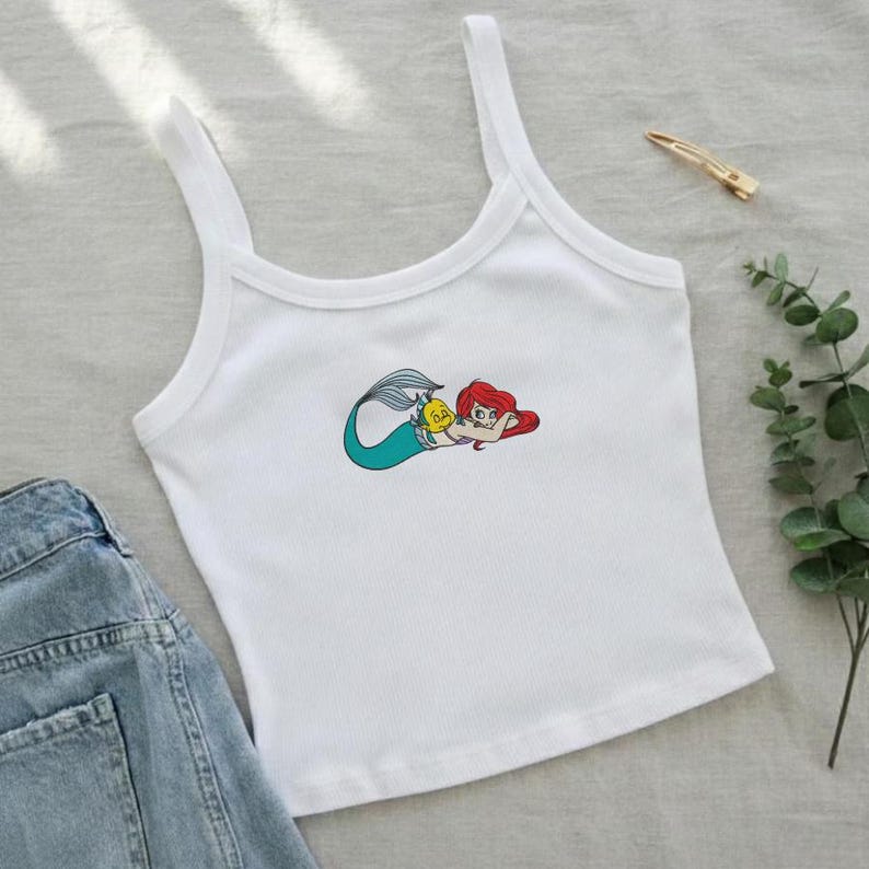 May include: White ribbed tank top with a colorful embroidered image of a mermaid with red hair and a turquoise tail. The mermaid is lying down with a yellow fish. A pair of denim shorts is visible in the image.