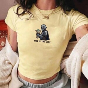 May include: Yellow crop top featuring a graphic of The Mandalorian holding Grogu, with the text "THIS IS THE WAY" in black. The shirt has short sleeves and a crew neck.