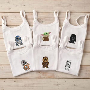 Pu&ograve; includere: Sei body bianchi per neonati con ricami di personaggi di Star Wars. I personaggi includono R2-D2, BB-8, Grogu, Chewbacca, Darth Vader e uno Stormtrooper. Ogni body ha un design senza maniche con spalline sottili.