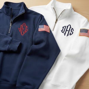May include: Two quarter-zip sweatshirts, one navy blue and one white. The navy sweatshirt has a red monogram and an American flag patch. The white sweatshirt has a navy monogram and an American flag patch. Both have a stand-up collar.