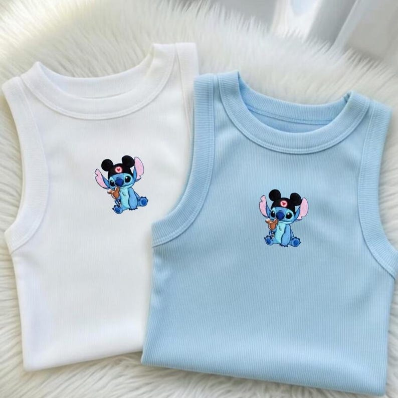 Puede incluir: Se muestran dos camisetas sin mangas, una blanca y otra azul claro. Cada camiseta presenta un personaje de dibujos animados bordado en el centro. El personaje es azul con orejas negras y un accesorio en forma de coraz&oacute;n. Las camisetas est&aacute;n hechas de tela suave y acanalada.