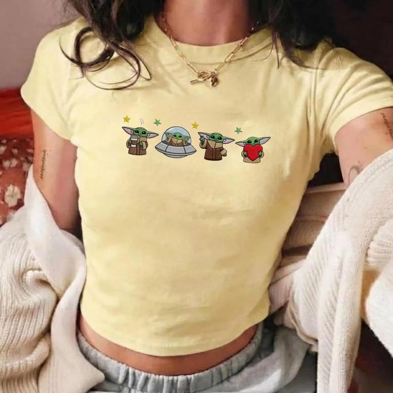 May include: A light yellow crop top featuring a cartoon design of a character from a popular science fiction series. The design includes four images of the character, with stars and a spaceship. The shirt has short sleeves.
