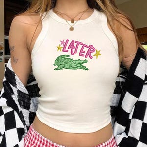 Peut inclure: Débardeur blanc avec un motif d'alligator vert et le mot "LATER" en rose avec des étoiles jaunes. Le haut est sans manches et a une encolure arrondie.