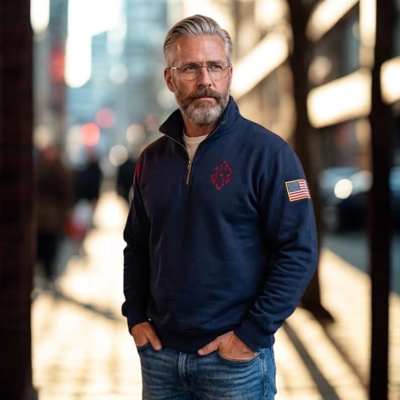 May include: Navy blue quarter-zip sweatshirt with a red embroidered emblem and an American flag patch on the sleeve. The sweatshirt has a stand-up collar and a silver zipper. The person is wearing glasses and has a gray beard.