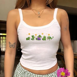 May include: White tank top with a scoop neck and spaghetti straps. The top features a row of green chameleon cartoon characters with yellow suns above them. The chameleons are in various poses, with one wearing a purple dress.