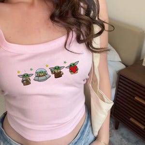 May include: A light pink tank top featuring a graphic of a green alien character with large ears. The graphic includes the alien in a spaceship, holding a cup, and holding a red heart. The tank top has a square neckline.