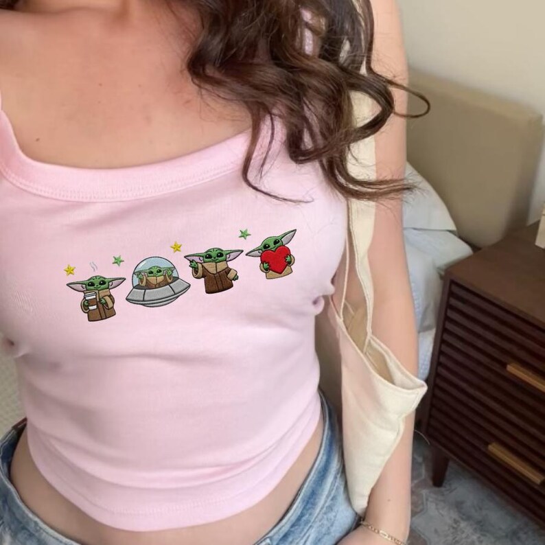 May include: A light pink tank top featuring a graphic of a green alien character with large ears. The graphic includes the alien in a spaceship, holding a cup, and holding a red heart. The tank top has a square neckline.