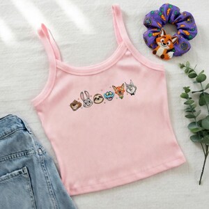 May include: Pink ribbed tank top with embroidered cartoon animal faces. A purple scrunchie with carrot and fox designs is next to the top. A pair of blue denim shorts is visible in the image.