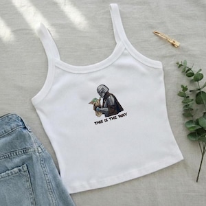 May include: White tank top with a graphic of a Mandalorian character holding Grogu, with the text "THIS IS THE WAY" in brown. The top has thin straps and a ribbed texture. A gold hair clip and greenery are also visible.