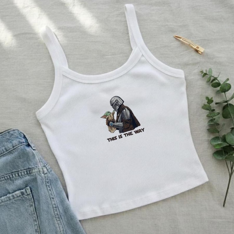 May include: White tank top with a graphic of a Mandalorian character holding Grogu, with the text "THIS IS THE WAY" in brown. The top has thin straps and a ribbed texture. A gold hair clip and greenery are also visible.