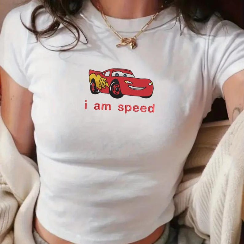 May include: White t-shirt featuring a red cartoon race car with the text "i am speed" in red. The car has yellow and red accents. The shirt is a short-sleeved, crew neck style.
