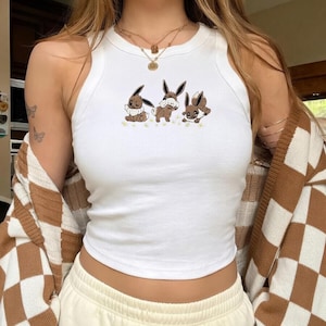 Puede incluir: Camiseta sin mangas blanca con cuello redondo con un diseño bordado de tres conejos de dibujos animados en marrón y blanco. Los conejos están colocados en fila con pequeñas flores amarillas. La camiseta se combina con pantalones deportivos color crema y una rebeca a cuadros marrón y blanco.