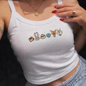May include: White tank top with embroidered cartoon animal faces. The design features a bear, rabbit, sloth, and other characters. The top has a scoop neck and thin straps.
