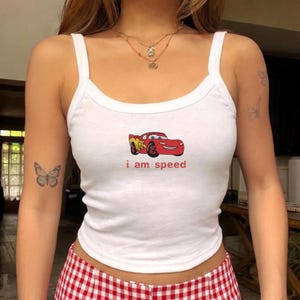 May include: White tank top with a red and yellow cartoon car graphic and the text "i am speed". The top has a scoop neck and thin straps. The model is wearing red and white checkered shorts.