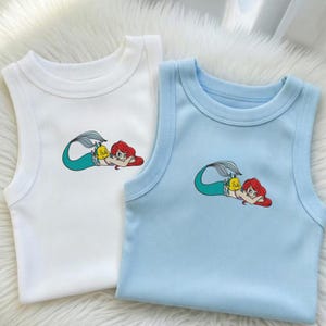 May include: Two sleeveless baby tank tops, one white and one light blue. Each top features a colorful cartoon mermaid graphic with red hair and a yellow fish. The tops are made of a soft, ribbed fabric.