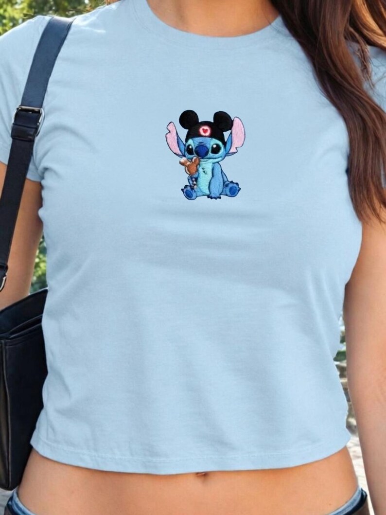 Puede incluir: Top corto azul claro con una imagen bordada del personaje de Disney Stitch. Stitch es azul, lleva un sombrero negro y sostiene un peque&ntilde;o osito de peluche marr&oacute;n. La parte superior es de color liso y tiene cuello redondo.