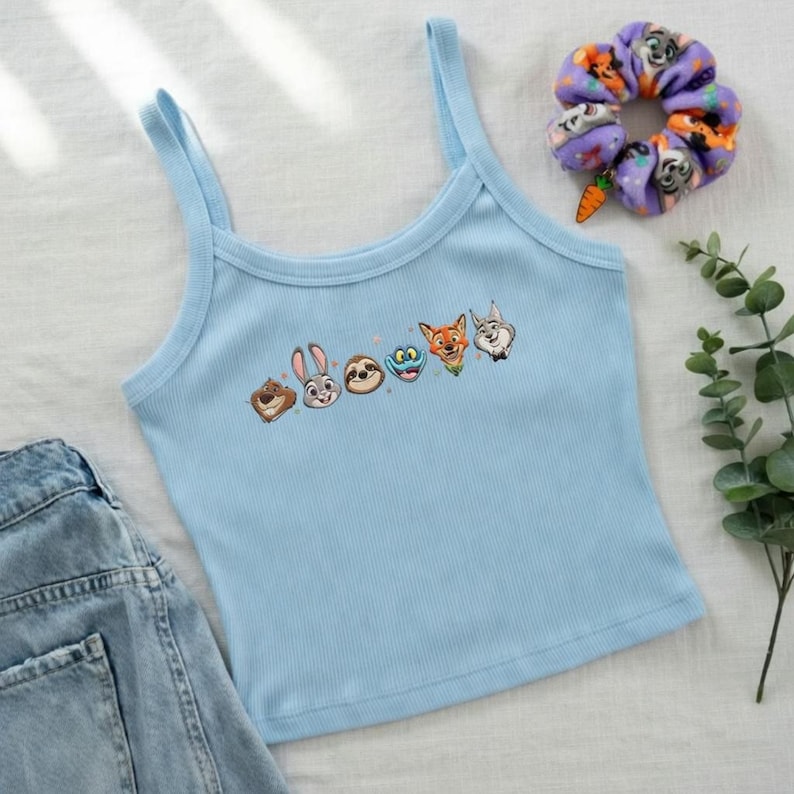 May include: Light blue tank top with a row of cartoon animal faces. A purple scrunchie with a carrot charm and a sprig of greenery are also in the image. The tank top is made of a ribbed material.