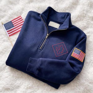 Könnte beinhalten: Marineblaues Sweatshirt mit Viertelreißverschluss, rotem gesticktem Logo und einem amerikanischen Flaggenaufnäher auf dem Ärmel. Das Sweatshirt hat einen gerippten Kragen und Bündchen. Eine kleine amerikanische Flagge ist ebenfalls im Bild.