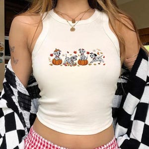 May include: A cream-colored sleeveless tank top featuring a graphic print of four spotted Dalmatian puppies interacting with orange pumpkins and scattered autumn leaves. The top has a ribbed crew neckline and a fitted silhouette.
