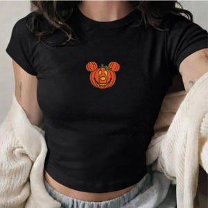 May include: A black short-sleeved t-shirt featuring a central graphic of an orange pumpkin shaped like a mouse head with a smiling face, a small green stem on top, and black outlines, set against a solid black fabric background.