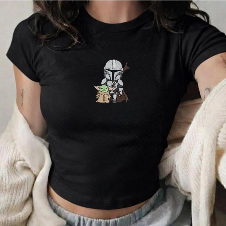 May include: Black crop top featuring an embroidered design of The Mandalorian and Grogu. The graphic shows the Mandalorian in silver armor and Grogu in a tan coat. The top has short sleeves and a crew neck.