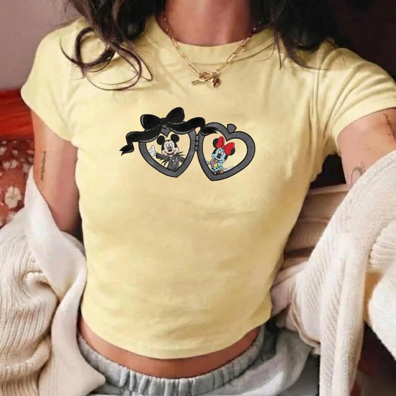 May include: A pale yellow short-sleeved t-shirt featuring a graphic of two heart-shaped lockets tied together with a black bow. Inside the left locket is a black and white Mickey Mouse, and inside the right is a colorful Minnie Mouse.
