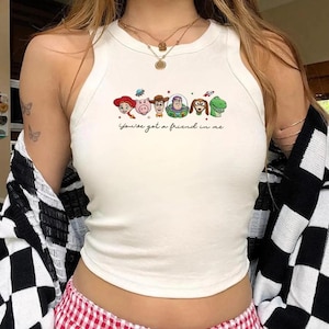 Peut inclure: D&eacute;bardeur blanc avec un graphisme color&eacute; de personnages anim&eacute;s et le texte "You've got a friend in me." Le motif comprend des personnages comme une cowgirl, un cochon, un cow-boy, un astronaute, un chien et un dinosaure. Le haut est sans manches.