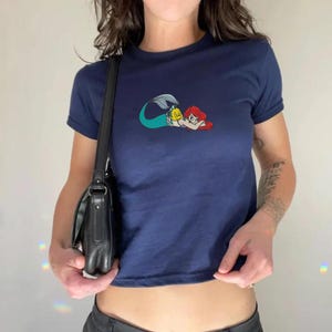 May include: Navy blue t-shirt featuring a colorful cartoon graphic of a mermaid with red hair and a yellow fish. The short-sleeved shirt is a cropped style.