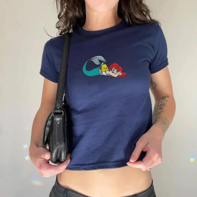 May include: Navy blue t-shirt featuring a colorful cartoon graphic of a mermaid with red hair and a yellow fish. The short-sleeved shirt is a cropped style.