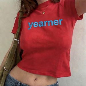 Op de afbeelding: Een rode crop top met het woord "yearner" in blauw geborduurd. De top heeft korte mouwen en een ronde halslijn. Een gouden ketting met een hartvormige hanger is zichtbaar. Het model draagt een blauwe jeans en een groene schoudertas.